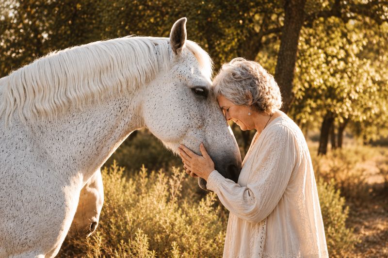 An older woman resting her forehead against a white horse in dappled forest light